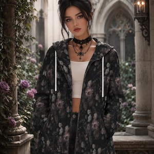 May include: A black zip-up hoodie featuring a dark floral pattern with muted purple and grey roses. The hoodie includes a hood with white drawstrings, long sleeves, and two front pockets. It is worn over a plain white crop top and dark jeans.