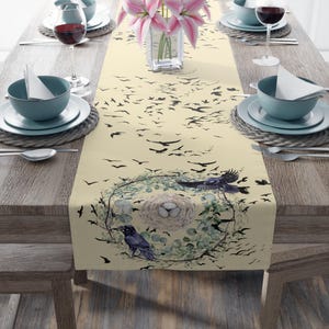 May include: A beige table runner with a black and white bird design. The runner features a wreath of green leaves and a bird's nest with eggs. The runner is on a wooden table with two place settings.