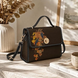 May include: A dark brown structured handbag featuring a top handle and a detachable shoulder strap. The front flap displays a printed design of two dark birds perched among vibrant orange and yellow autumn leaves, secured by a gold metal turn-lock.