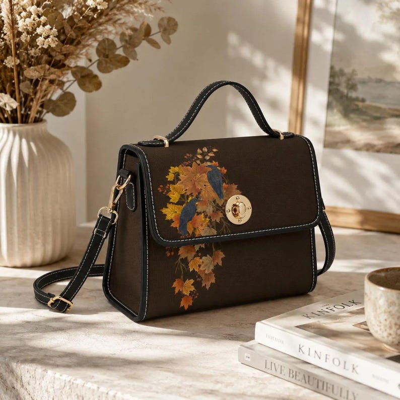 May include: A dark brown structured handbag featuring a top handle and a detachable shoulder strap. The front flap displays a printed design of two dark birds perched among vibrant orange and yellow autumn leaves, secured by a gold metal turn-lock.