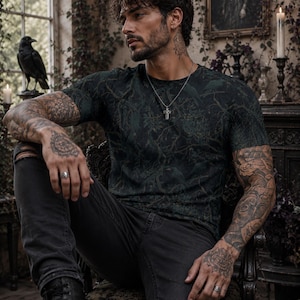 Pode incluir: Uma t-shirt de manga curta verde-escura com um padr&atilde;o subtil de floresta e p&aacute;ssaros, combinada com cal&ccedil;as de ganga pretas desgastadas e botas de couro pretas. O conjunto inclui um colar com pendente de cruz e an&eacute;is de prata nos dedos.