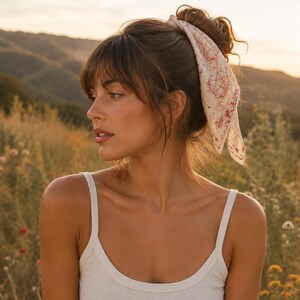 May include: A square hair scarf featuring a cream-colored base with a repeating red print of celestial symbols, including a pentagram, crescent moons, and stars, tied around a high bun hairstyle with loose bangs framing the face.