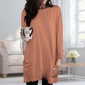 May include: A long-sleeved, solid terracotta-colored tunic top featuring a round neckline and two large patch pockets on the front. The garment has a relaxed, loose-fitting silhouette that extends past the hips, providing a comfortable and casual look.