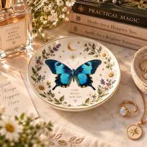 May include: A round white ceramic trinket dish featuring a large blue butterfly at the center, surrounded by floral illustrations of daisies and lavender. The dish includes a gold crescent moon, stars, and the text SAME MAGIC NEW DAY with a heart.