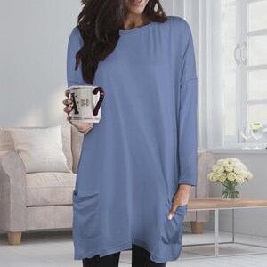 May include: A long-sleeved, periwinkle blue tunic top featuring a round neckline, a relaxed fit, and two large patch pockets on the front. The garment is made of a solid-colored fabric and extends to the mid-thigh, providing a simple and clean silhouette.