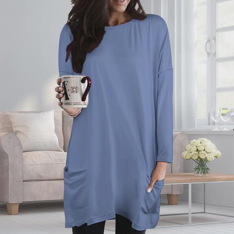 May include: A long-sleeved, periwinkle blue tunic top featuring a round neckline, a relaxed fit, and two large patch pockets on the front. The garment is made of a solid-colored fabric and extends to the mid-thigh, providing a simple and clean silhouette.