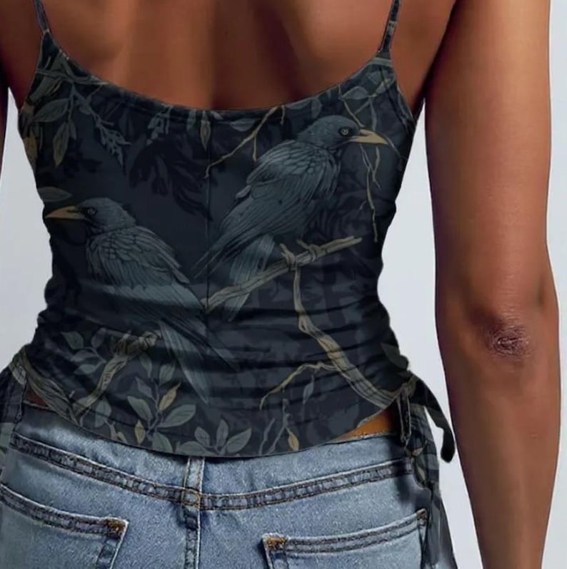 May include: A dark-toned camisole featuring a repeating pattern of black crows perched on branches amidst foliage. The garment has thin spaghetti straps and a side-tie detail at the hem, worn over light blue denim jeans with visible pockets.