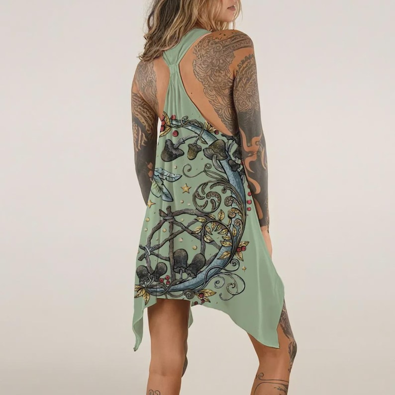 May include: A sage green racerback tank top featuring a graphic print of a crescent moon, swirling vines, small stars, and various mushrooms. The garment has an asymmetrical hemline and a loose, flowing fit that hangs from the shoulders.