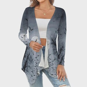 May include: A long-sleeve open-front cardigan featuring a gray and white graphic print of a moonlit forest scene with silhouettes of flying bats. The garment has a draped hemline and is worn over a plain white crop top and blue denim jeans.