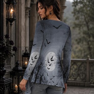 May include: A long-sleeved tunic featuring a gray-toned print of a full moon, silhouetted bare trees, and a flock of flying bats. The garment has a rounded neckline and an asymmetrical hemline, paired with fitted gray denim jeans.