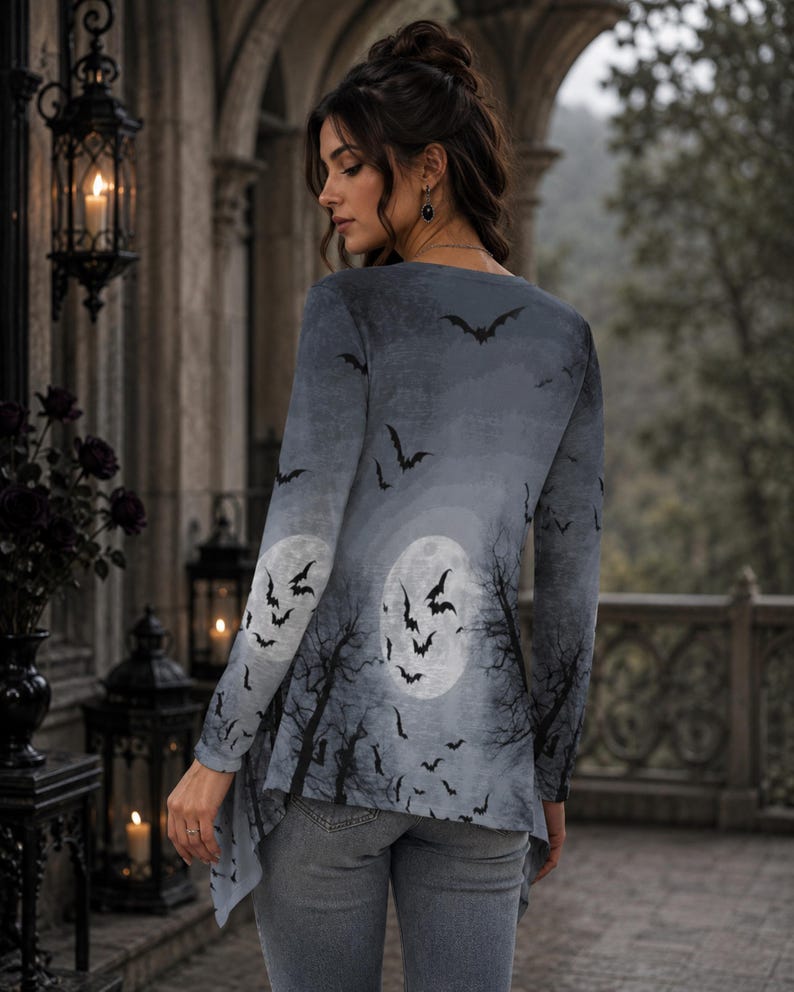 May include: A long-sleeved tunic featuring a gray-toned print of a full moon, silhouetted bare trees, and a flock of flying bats. The garment has a rounded neckline and an asymmetrical hemline, paired with fitted gray denim jeans.