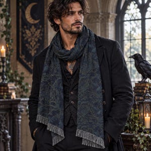 May include: A dark blue scarf with a subtle, intricate pattern of branches and birds, featuring a fringed hem. The scarf is worn over a black button-down shirt, a dark vest, and a black coat, with a silver pendant necklace visible at the neckline.