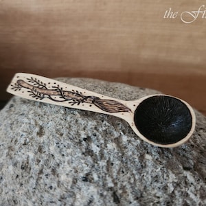 Könnte beinhalten: Ein kleiner Holzlöffel mit einem schwarzen, eingebrannten Design auf der Schale und einem fantasievollen Design aus einem Besen und Ranken, das in den Griff eingebrannt ist. Der Löffel liegt auf einem grauen Stein.
