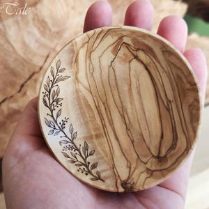 Könnte beinhalten: Eine kleine, runde Holzschale mit einem dekorativen Blatt- und Beerenmuster. Die Schale besteht aus hellbraunem Holz mit einem natürlichen Maserungsmuster. Das Blattmuster ist dunkelbraun und befindet sich auf der linken Seite der Schale.