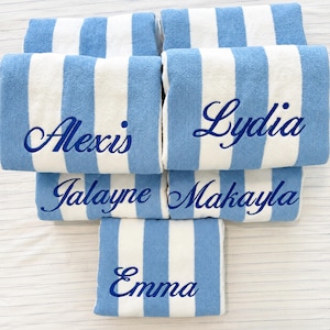May include: Five folded rectangular beach towels featuring vertical light blue and white stripes. Each towel is embroidered with a name in dark blue cursive script: Alexis, Lydia, Jalayne, Makayla, and Emma. The towels are arranged in a stack.