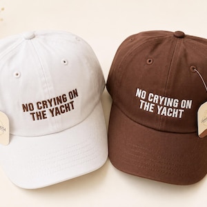May include: Two baseball caps, one white and one brown, each featuring the embroidered text "NO CRYING ON THE YACHT" on the front. The white cap has brown lettering, while the brown cap has white lettering. Both caps include a small brand tag.