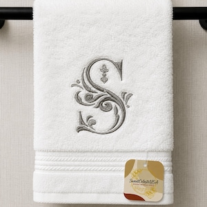 May include: A white terry cloth towel featuring a large, gray embroidered letter S with ornate, swirling filigree details. The towel has a textured border at the bottom and includes a small, square tag with the text SweetCelesteUSA.
