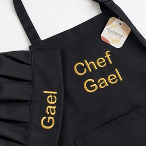 Może przedstawiać: Czarny fartuch i pasująca czapka kucharska z wyhaftowanym złotym napisem. Na fartuchu widnieje napis Chef Gael, a na czapce imię Gael. Do paska fartucha przymocowana jest metka marki z napisem SweetCelesteUSA.