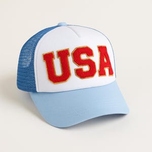 Pode incluir: Boné de beisebol com painel frontal branco e aba azul claro. A palavra "USA" é bordada em vermelho com acabamento dourado na frente. A parte de trás do boné é de malha azul.