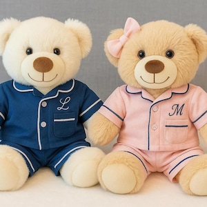 May include: Two plush teddy bears wearing matching pyjamas. One bear is cream-coloured and wears navy blue pyjamas with a white trim and the letter "L". The other bear is tan and wears pink pyjamas with a navy trim and a pink bow.