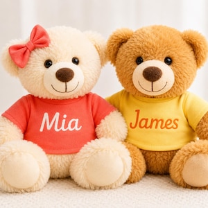 Puede incluir: Dos osos de peluche sentados uno al lado del otro. El oso color crema de la izquierda lleva un lazo rojo y una camiseta roja con el nombre Mia impreso en blanco. El oso marrón de la derecha lleva una camiseta amarilla con el nombre James en naranja.