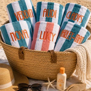 May include: A woven straw basket filled with six folded beach towels featuring teal and white or coral and white stripes. Each towel is embroidered with orange text reading Verdin, Audi, Cladys, Nova, Lucy, or Harley-Man. A hat, sunglasses, sunscreen, and a starfish are nearby.