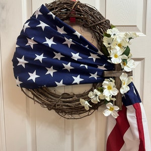May include: A patriotic wreath decorated with a red, white, and blue American flag, white flowers, and a wooden cross.