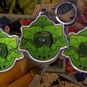 May include: Three clear acrylic keychains with a green leaf design. Each keychain has a small, colorful character inside. The keychains are attached to silver keyrings. The background includes seed packets and produce.
