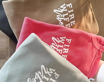 Pode incluir: Quatro moletons em cores diferentes, empilhados uns sobre os outros. Os moletons são todos do mesmo estilo e têm o texto "FIRE WIFE VIBES" impresso em branco.
