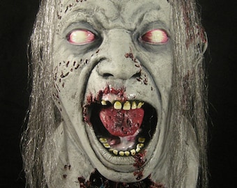 May include: A realistic zombie mask with a pale gray face, long white hair, and a wide open mouth revealing bloody teeth and a red tongue. The mask has red blood splatters and blue veins on the neck.