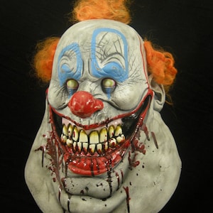 May include: A creepy clown mask with a wide, toothy grin and blood dripping from its mouth. The mask is made of latex and has a realistic appearance. The clown has a white face with blue and red makeup, and orange hair.