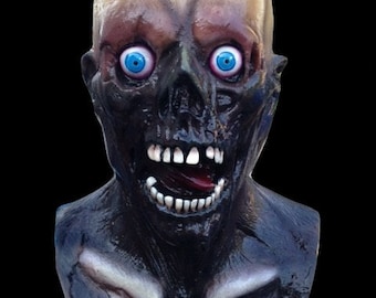 Può includere: Una grottesca maschera da zombie con un cranio giallo pallido, lineamenti scuri neri e occhi blu brillante. La maschera ha la bocca aperta che rivela denti bianchi e una lingua rossa. L'area del collo è blu scuro con accenti bianchi, su sfondo nero.