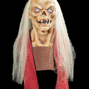 May include: A white-haired, pale-skinned mask with a grotesque expression. The mask has large, blue eyes and a wide, toothy grin. The mask is wearing a red scarf.