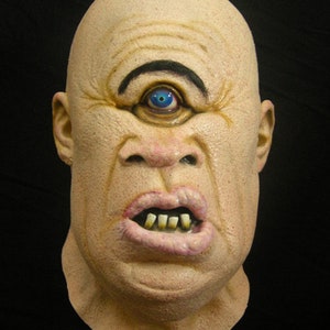 May include: A close-up of a grotesque, pale-skinned mask with a single, large blue eye and a wide, toothy grin. The mask is made of a textured material and appears to be a Halloween costume.