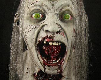May include: A gruesome zombie mask with a pale complexion, green eyes, and exposed teeth. The mask features open wounds and a bloody mouth, with long gray hair. A horror-themed costume accessory.
