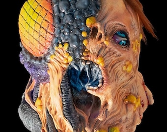 May include: A grotesque, multi-colored mask with a half-human, half-insectoid design. The mask features a large, segmented eye in yellow and orange, juxtaposed with decaying flesh tones, blue and black textures, and exposed teeth. The overall theme is horror.