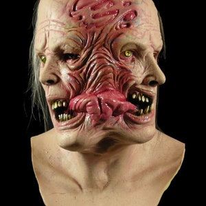 Könnte beinhalten: Eine groteske, realistische Horror-Maske mit einem fleischigen, entstellten Gesicht. Die Maske hat zwei Sätze Zähne und Augen, mit einem aufgerissenen Mund und einer fleischigen, rosa Zunge.