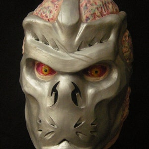 Op de afbeelding: Een gedetailleerd masker in zilver en rood met een schedelontwerp en rode ogen. Het masker is gemaakt van een hard materiaal en heeft een textuur.