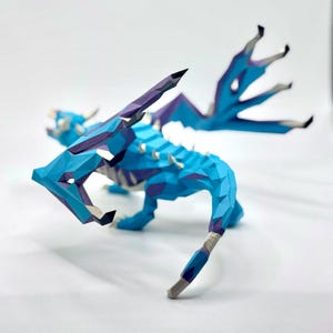 May include: A geometric paper dragon sculpture in shades of blue, purple, and white. The dragon has detailed wings and a spiky spine. The artwork is set against a white background, highlighting the intricate papercraft design.