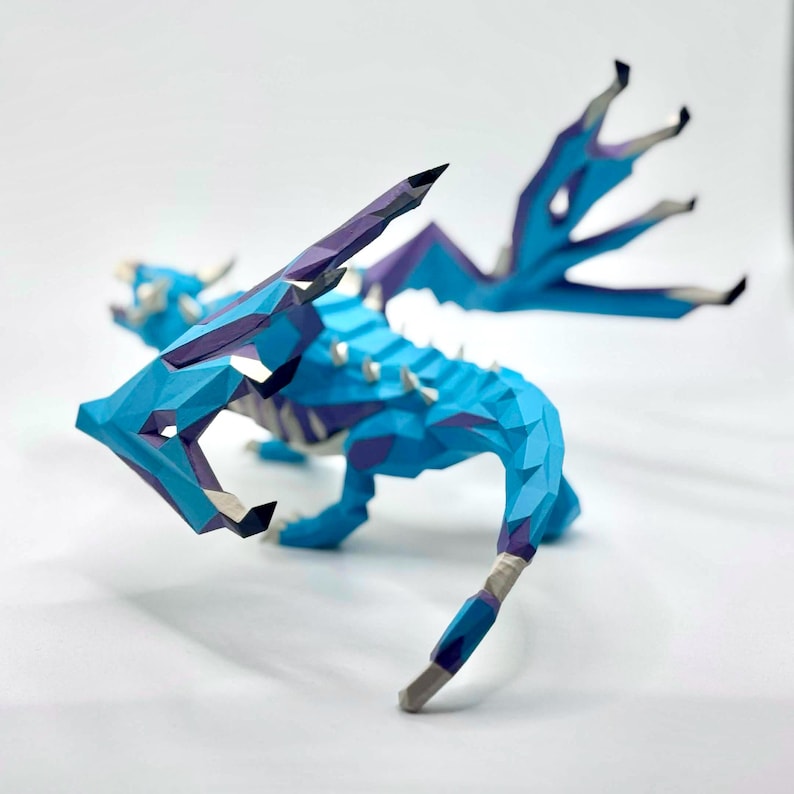 May include: A geometric paper dragon sculpture in shades of blue, purple, and white. The dragon has detailed wings and a spiky spine. The artwork is set against a white background, highlighting the intricate papercraft design.
