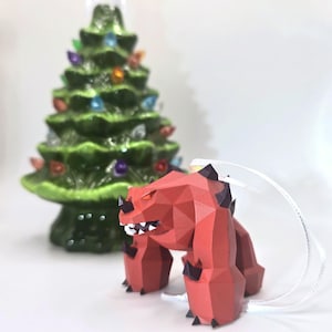 May include: Red and black polygonal Christmas ornament shaped like a monster with a toothy grin. The ornament is hanging from a white string.