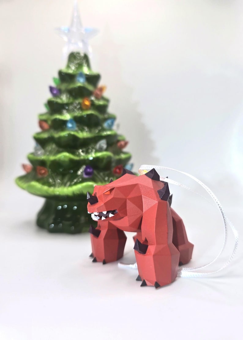 May include: Red and black polygonal Christmas ornament shaped like a monster with a toothy grin. The ornament is hanging from a white string.