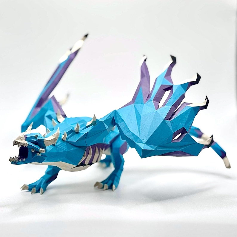 May include: A blue and purple papercraft dragon with open mouth and outstretched wings. The dragon has a geometric design with sharp angles and a detailed skeletal structure. The wings are a mix of blue and purple, and the dragon has white horns and teeth.