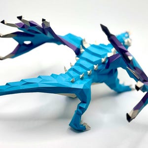 May include: A low-poly, geometric dragon sculpture in shades of blue, purple, and white. The dragon has sharp, white spikes along its back and wings. The wings are spread, and the dragon is posed as if in motion.