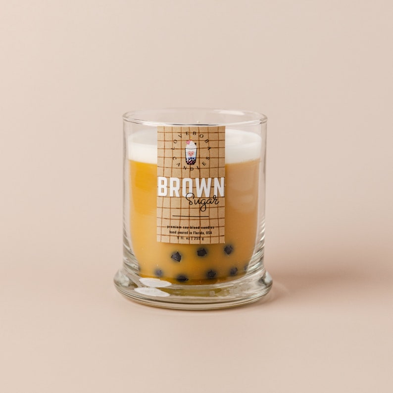 May include: A glass jar candle with a brown sugar scent. The candle is topped with a layer of white wax and has a label that says "Brown Sugar" and features a boba tea graphic.