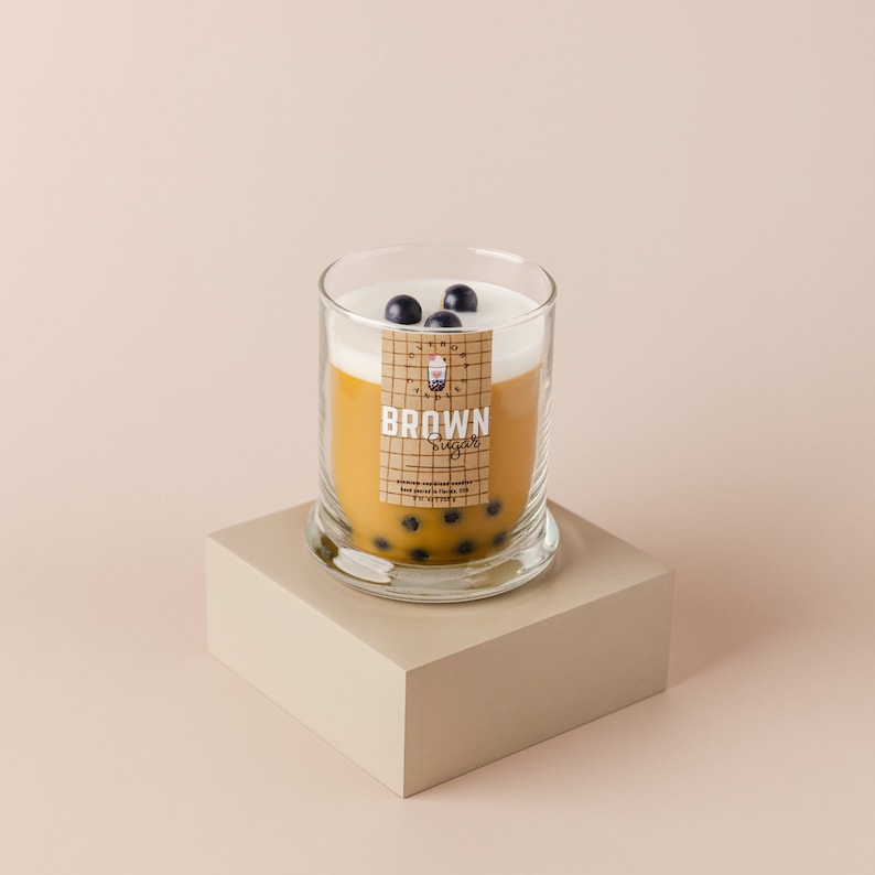 May include: A glass candle jar with a brown sugar scented candle. The candle is topped with white wax and black boba pearls. The label on the jar reads "BROWN Sugar" with a cartoon image of a boba tea.