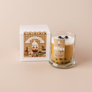 May include: A brown sugar boba tea scented candle in a clear glass jar with a brown sugar boba tea illustration on the label. The candle is topped with a white wax layer and blue boba pearls. The label reads "LOVE BOBA CANDLES BROWN Sugar".