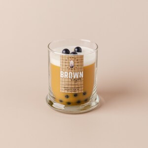 May include: A glass jar candle with a brown sugar scent. The candle is topped with a white layer of wax and black boba pearls. The label reads "BROWN Sugar" and features a cartoon boba tea cup.
