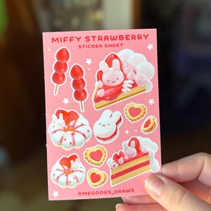 May include: A pink sticker sheet featuring illustrations of a bunny character and strawberry-themed treats. The sheet includes images of strawberry skewers, cake slices, and heart-shaped cookies. Text on the sheet reads "MIFFY STRAWBERRY STICKER SHEET" and "@MEGOOGS_DRAWS".