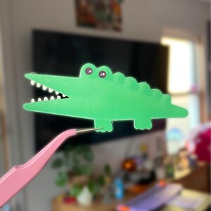 May include: A green paper alligator with a toothy grin and googly eyes is held up by a pair of pink tweezers.